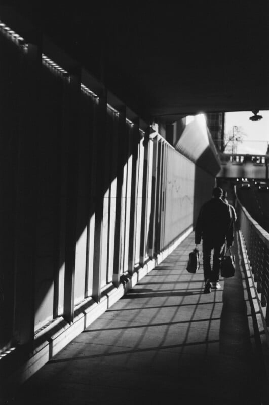 A person holding bags walks through a shadowy, sunlit tunnel with geometric shadows cast on the ground and walls.