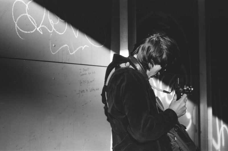 A person in a dark jacket stands under a beam of light, looking down at a film camera. Graffiti and handwritten notes are visible on the wall beside them. The scene appears to be indoors and in black and white.