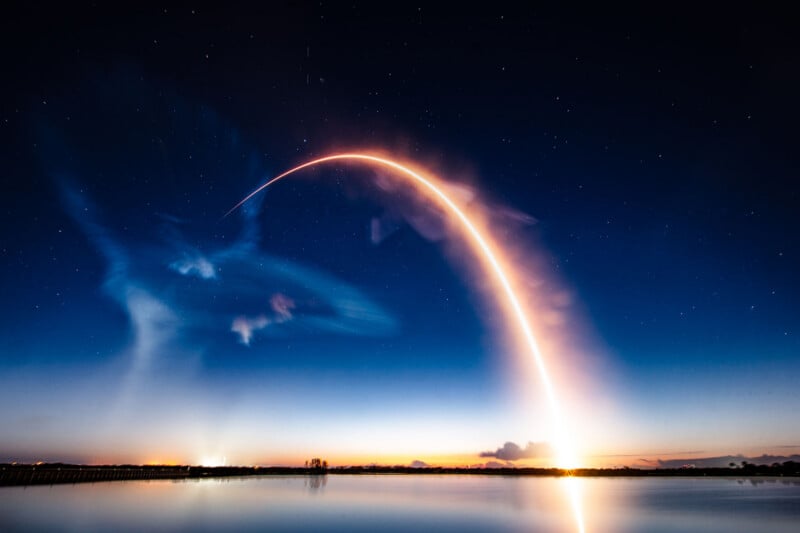 A long-exposure photo of a rocket launch creating a bright arc across a twilight sky, reflected in calm water, with stars visible and wispy clouds illuminated by the rocket's trail.