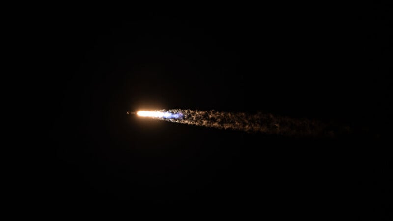 A bright rocket soars upward against a dark night sky, leaving a glowing trail of exhaust and smoke behind it.