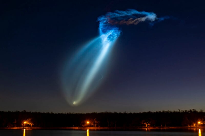 A bright, glowing blue and white light trail arcs across the night sky, with illuminated clouds above and a dark tree line and lights reflected in a calm lake below.