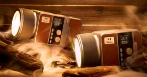 Two retro-styled, brown and silver devices with dials and screens sit among pieces of wood and swirling mist, illuminated warmly. They resemble vintage cameras but function as modern electronic gadgets.
