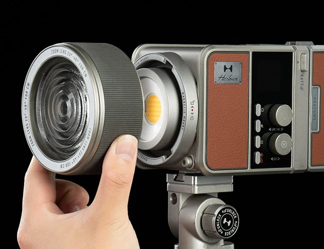 A hand attaches a large lens to a modern, retro-styled digital camera mounted on a tripod, against a black background.