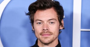 A young white man with wavy dark brown hair, green eyes, and light stubble wearing a black top, posed in front of a blue backdrop.