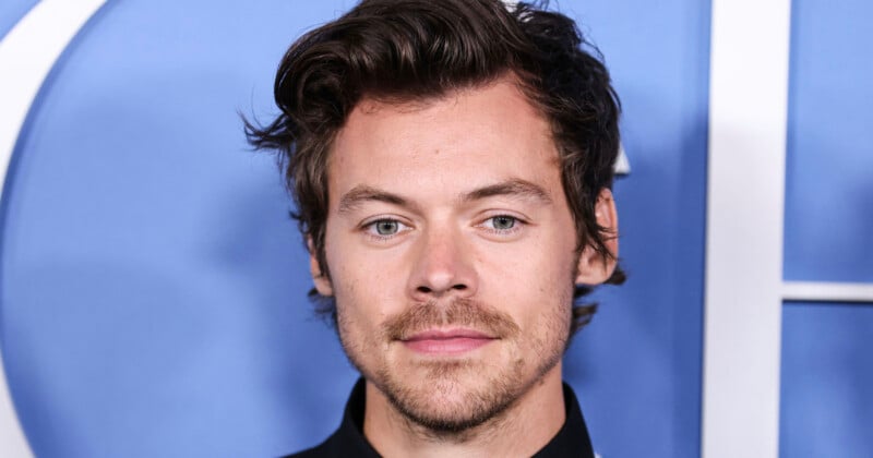 A young white man with wavy dark brown hair, green eyes, and light stubble wearing a black top, posed in front of a blue backdrop.