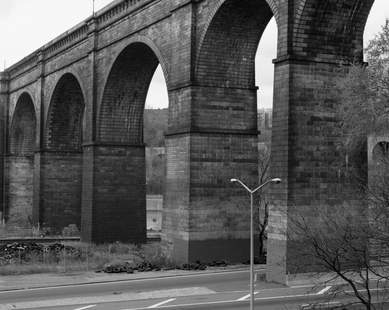 Una fotografía en blanco y negro de un gran viaducto ferroviario de piedra con altos arcos que cruzan la carretera. Había montones de neumáticos apilados cerca de la base y en primer plano se podían ver árboles desnudos.