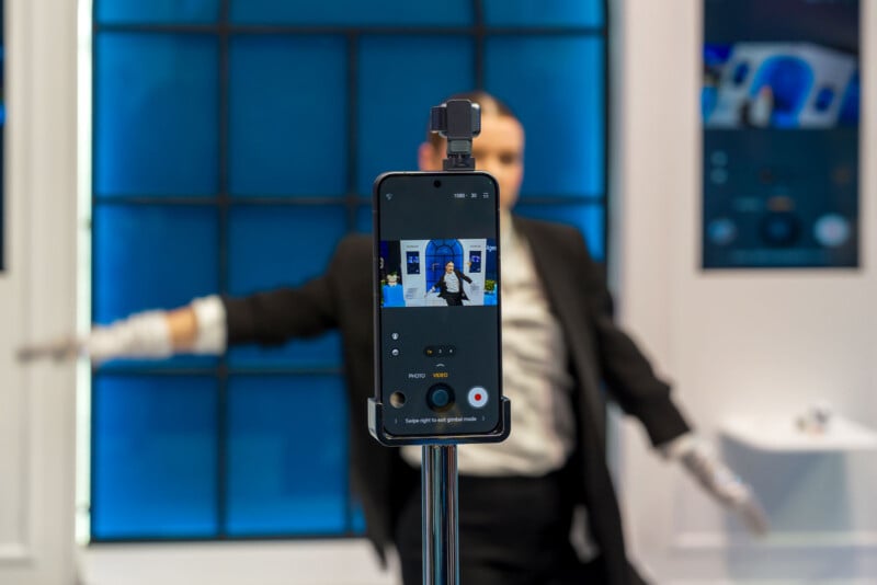 A smartphone on a tripod records a person in a suit and gloves posing with arms outstretched in front of a blue window. The person's image is visible on the phone screen.