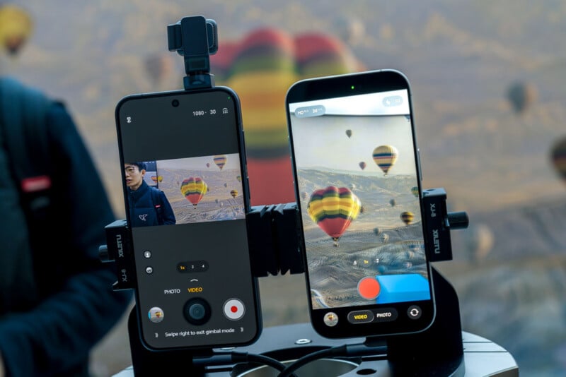 Two smartphones on a dual mount record and photograph colorful hot air balloons floating over a scenic, mountainous landscape. Both screens display the balloons from different camera modes.