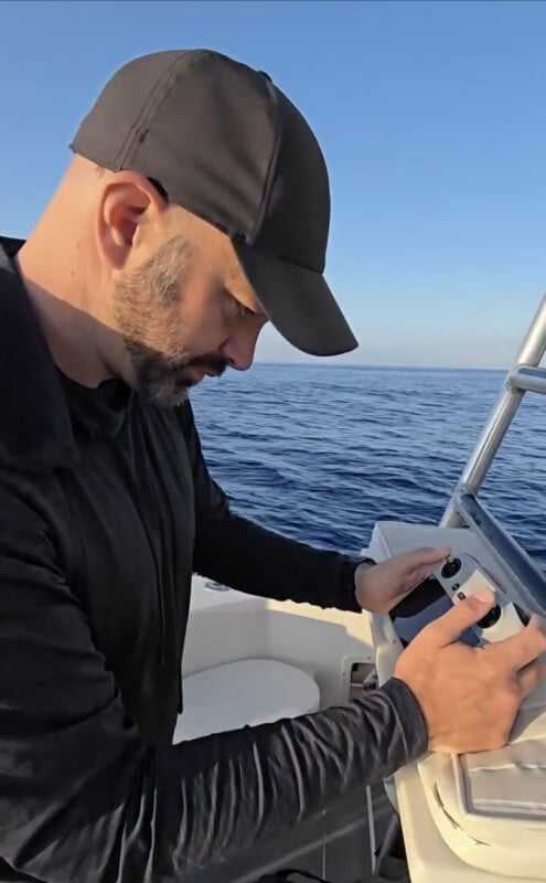 A man with a beard and black cap operates equipment on a boat, with calm blue ocean and clear sky in the background.
