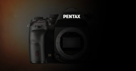 A digital camera body labeled "Pentax" is shown in dim lighting, with the lens removed, revealing the sensor and inner mount. The left side and background are dark, fading into black.
