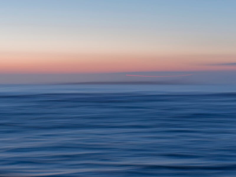 A blurred photo of the ocean at sunset, with soft waves in the foreground and a pastel-colored sky in shades of orange, pink, and blue. A faint, curved red line appears near the horizon.