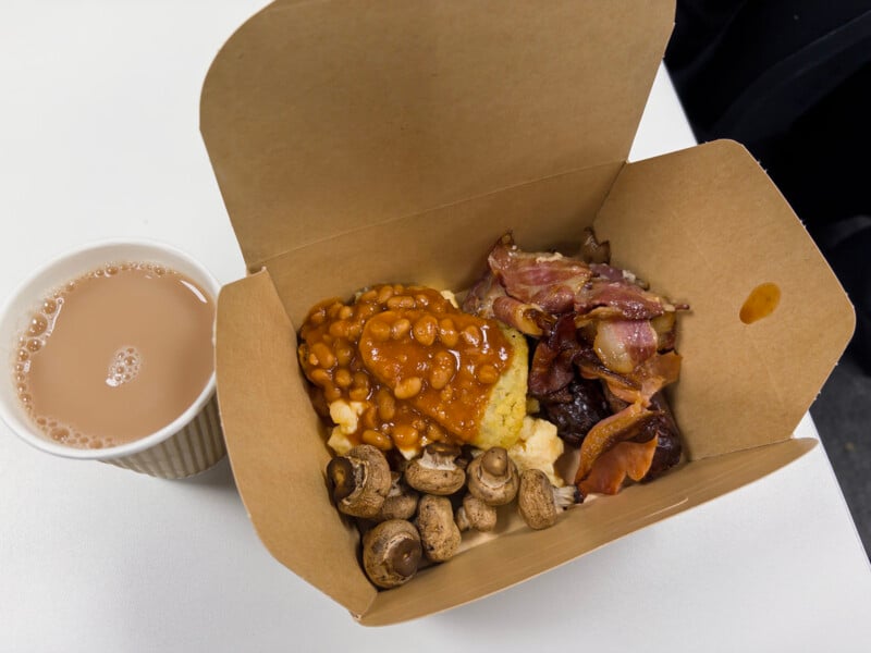 A cardboard takeout box contains baked beans on a hash brown, scrambled eggs, grilled mushrooms, and crispy bacon. Next to it is a cup of tea with milk on a white table.