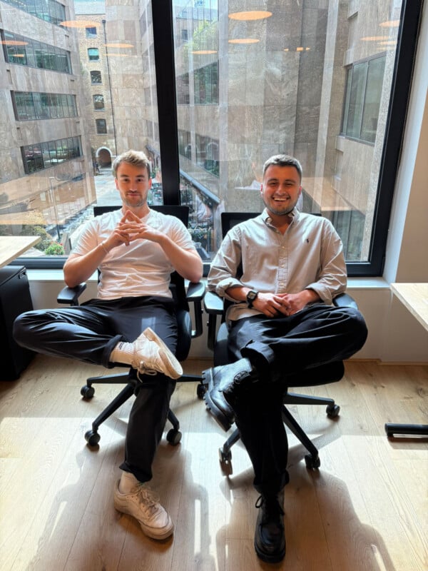 Dos hombres estaban sentados casualmente en sillas de oficina junto al gran ventanal, sonriendo a la cámara. La luz del sol entra a raudales, resaltando el moderno entorno de oficinas, y los edificios exteriores son claramente visibles. Los dos parecían relajados con las piernas cruzadas.