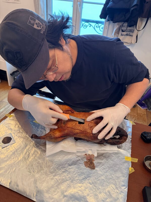 A person wearing glasses, a cap, and gloves uses a tool to carve a roasted duck on a table covered with foil, with a window and coats in the background.