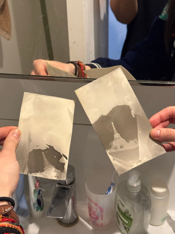 Two hands hold faded black and white photos in front of a bathroom mirror above a sink with soap dispensers. One photo shows the Eiffel Tower, and a reflection of the scene is visible in the mirror.