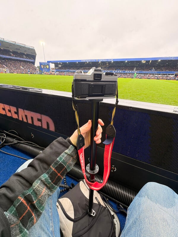 A person sitting pitchside at a football stadium holds a camera on a monopod, with the field and crowded stands visible in the background under an overcast sky.