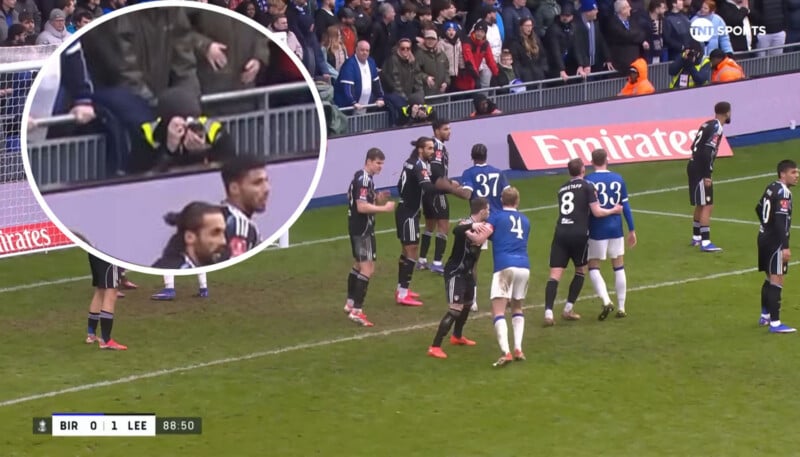 A soccer match between Birmingham (BIR) and Leeds (LEE) with players preparing for a set piece. Inset zooms in on a fan sitting on the barrier with his foot caught between metal bars.