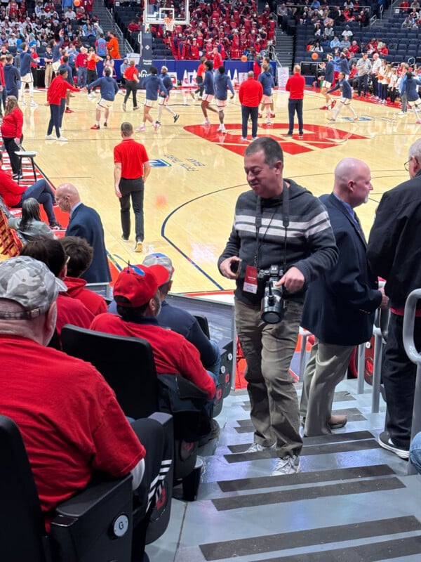 Un fotógrafo estaba en las escaleras de la arena con una cámara, hablando con los fanáticos sentados. Los espectadores con camisetas rojas llenaron los asientos y, al fondo, los jugadores de baloncesto calentaban en la cancha.