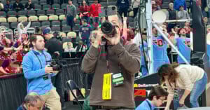 A photographer aims a camera at the viewer on a basketball court, with a sports crowd and band cheering in the background. Other media personnel and event staff are also visible around him.