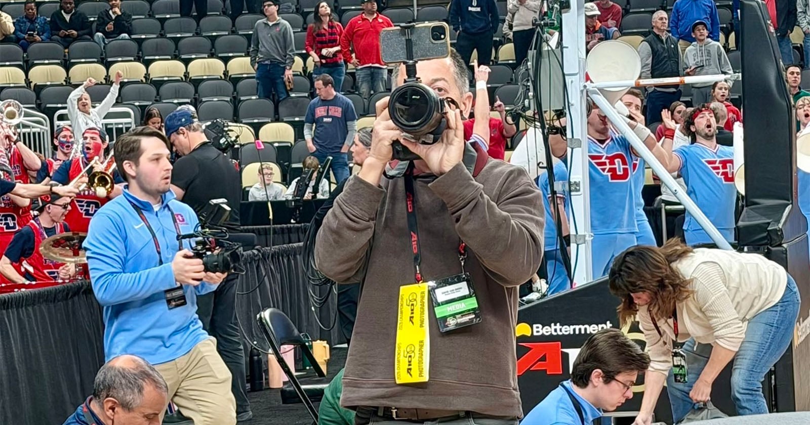 A photographer aims a camera at the viewer on a basketball court, with a sports crowd and band cheering in the background. Other media personnel and event staff are also visible around him.