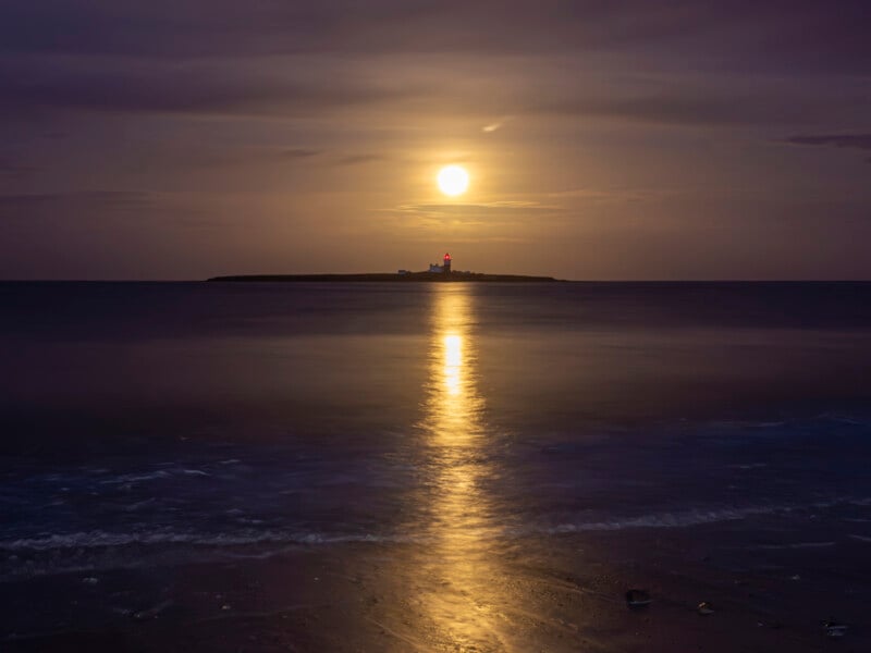 A full moon rises over a calm sea, casting a golden reflection on the water. In the distance, a small island features a lighthouse with its light shining faintly in the dusk.