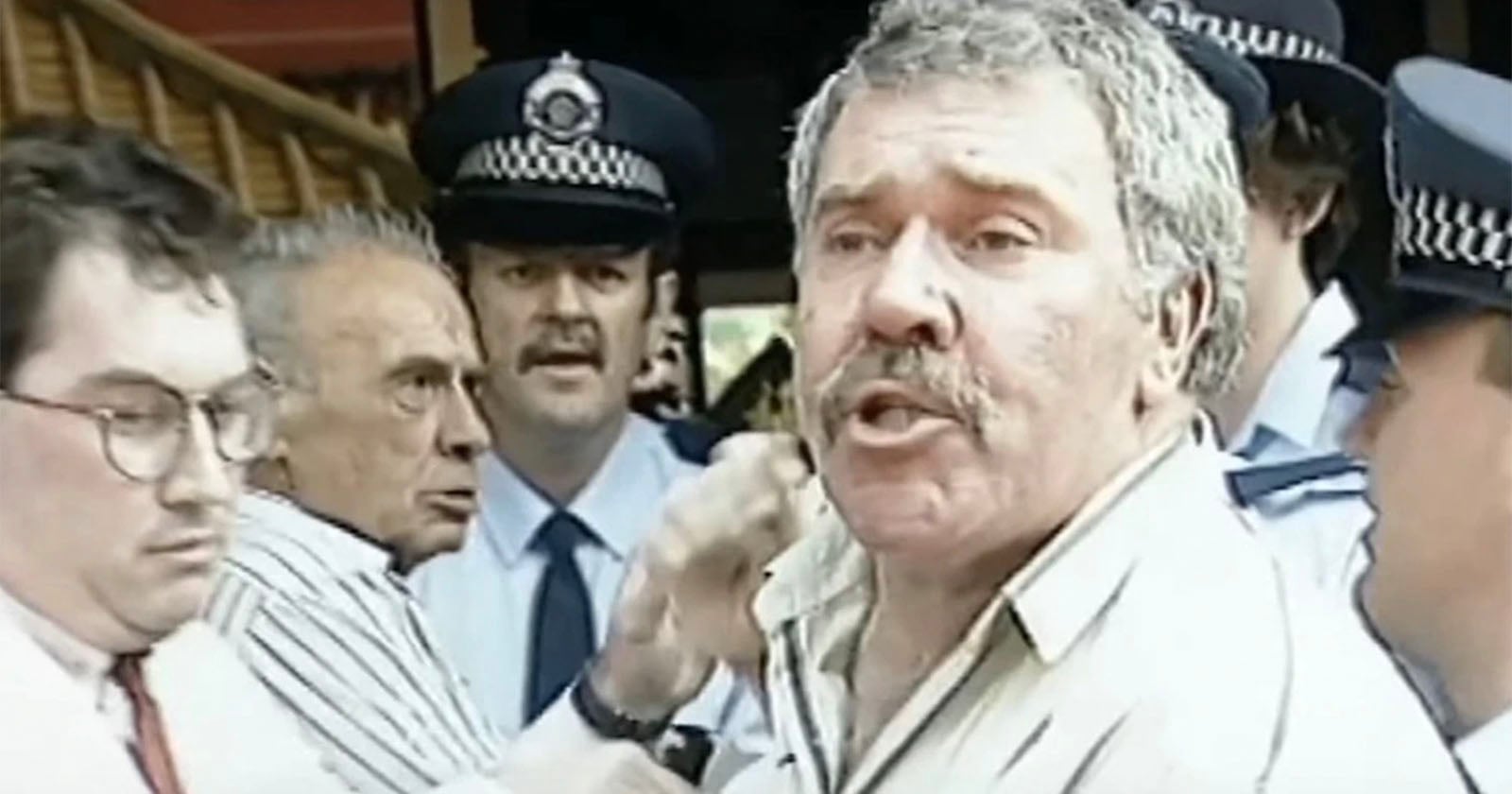 A man with a mustache is being held by police officers in uniform and another man in glasses. The scene appears tense, with several faces showing concern or urgency. The background includes a staircase and people.