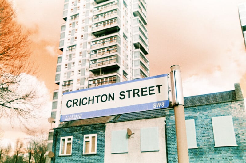 A street sign reading "CRICHTON STREET" stands in front of a blue brick building, with a tall modern apartment building and an orange-tinted sky in the background.