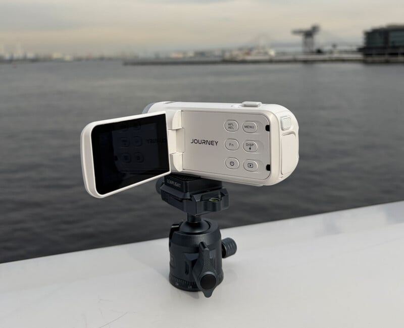 A white handheld video camera marked "JOURNEY" sits on a small tripod, facing water with an industrial shoreline and cloudy sky in the background. The camera's flip-out screen displays its control menu.