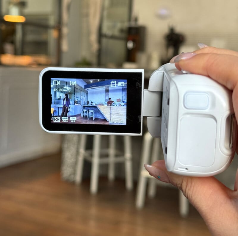 A hand holds a white video camera, focused on its screen, which displays a recording of a modern cafe with people sitting at tables inside. The background shows a blurred view of the same cafe.
