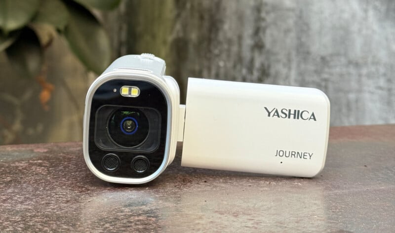 A close-up of a white Yashica Journey digital camcorder resting on a surface, with its flip-out screen open and the camera lens facing forward. The background is blurred with some greenery visible.