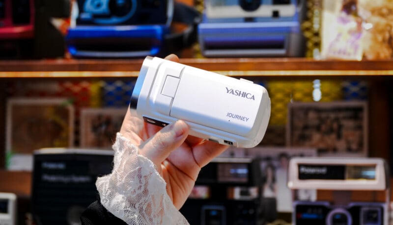 A person with lace-trimmed clothing holds a white Yashica Journey camera in their hand, with a background of vintage cameras and colorful decor on shelves.