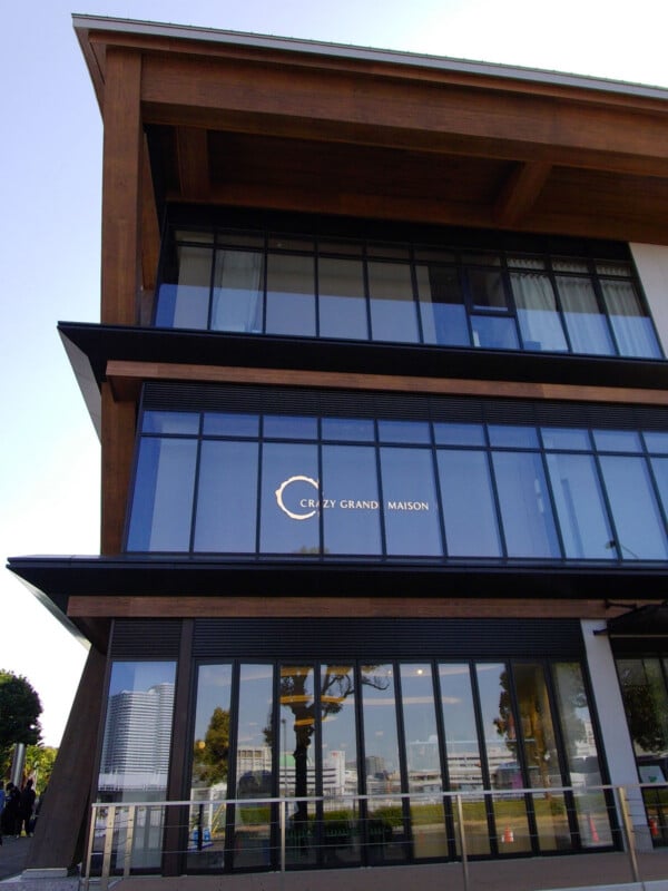 A modern, multi-story building with large glass windows and wooden beams. The sign "CRAZY GRAND MAISON" is visible on the second floor. Trees and city reflections are seen in the glass.