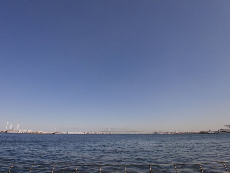 A wide view of a calm harbor with blue water and a clear sky; distant industrial buildings, wind turbines, and a bridge are visible on the horizon.