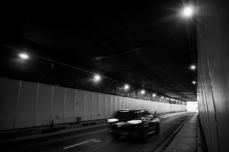 Una fotografía en blanco y negro muestra un automóvil acelerando a través de un túnel poco iluminado con luces brillantes en el techo y una acera vacía a la derecha. Hay luz al final del túnel.