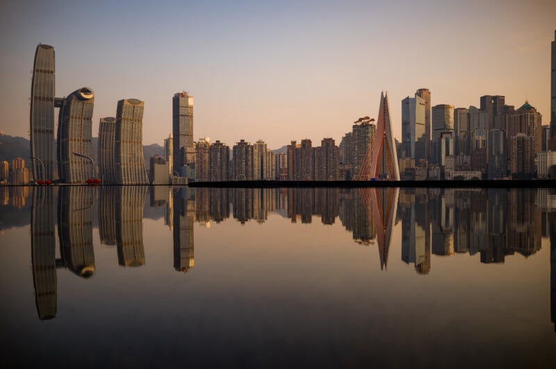 Al atardecer, el horizonte de la ciudad bordeado de rascacielos se refleja en el agua tranquila, creando un efecto simétrico similar a un espejo. El cielo estaba despejado y tenía un cálido tono dorado.