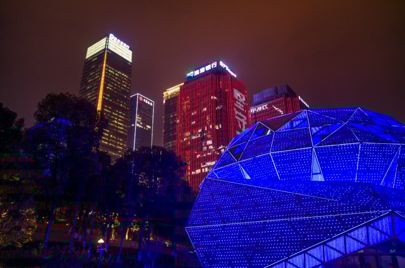En primer plano hay una cúpula geodésica de color azul brillante, con rascacielos rojos y amarillos brillantes que se elevan sobre un cielo nocturno nublado al fondo. Los árboles se pueden ver en el lado izquierdo de la imagen.