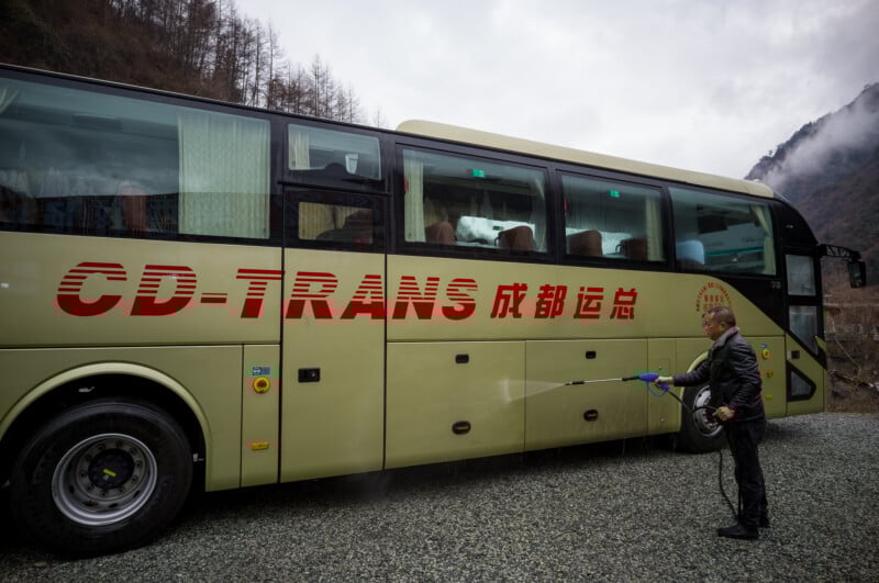 Un hombre rocía agua para limpiar el costado de un gran autobús color crema con etiquetas "CD-TRANS" El coche con caracteres chinos está aparcado sobre una superficie de grava cerca de la montaña.
