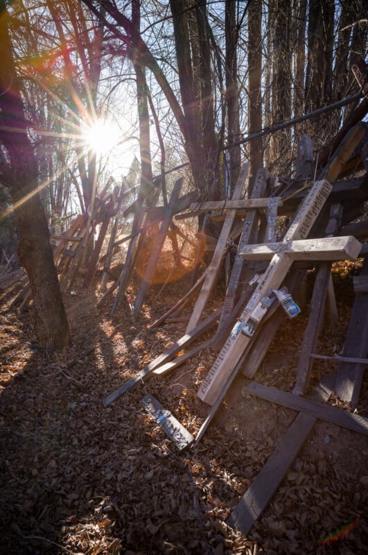 Una cruz de madera se alza en un sendero forestal soleado y cubierto de hojas. El sol brilla a través de las ramas desnudas, proyectando largas sombras e iluminando el campo.