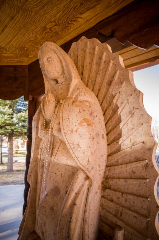 Una estatua de piedra de la Virgen María con las manos juntas y un rosario en la cabeza se encuentra bajo un techo de madera, con las figuras y los árboles claramente visibles al fondo bajo la luz del sol.