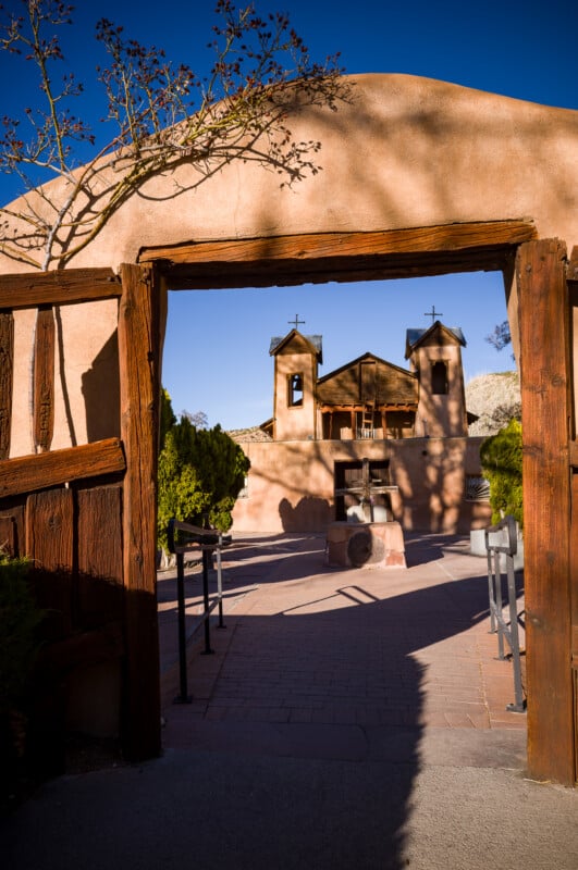 Un arco de adobe enmarca una vista de una iglesia histórica con dos campanarios contra un cielo azul claro. La luz del sol proyecta sombras de árboles en las paredes y pisos, y el camino de entrada está bordeado de árboles verdes.