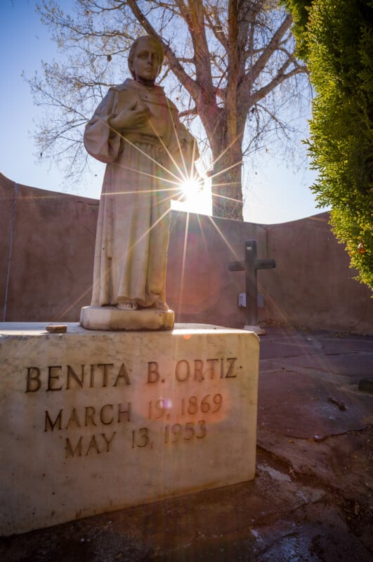 La lápida de mármol de Benita B. Ortiz (1869-1953) presenta una estatua de la santa, con la luz del sol brillando detrás de la estatua y un árbol y una cruz visibles al fondo.
