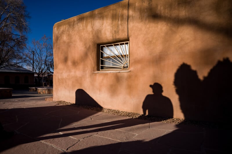 Bajo el cielo azul, una figura con sombrero se proyecta sobre una casa de adobe con una verja de hierro. Al fondo se ven claramente árboles desnudos y elementos del patio.