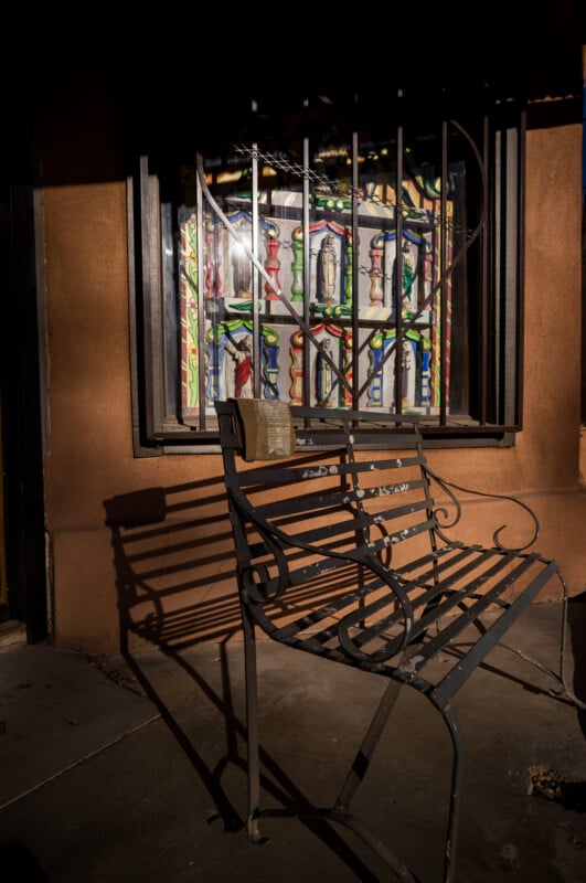 Un banco de metal proyecta una sombra en la acera frente a la ventana, que presenta una barra abstracta colorida y una reja de seguridad en forma de corazón. La luz del sol incide sobre los bancos y algunas de las ventanas, creando un fuerte contraste.