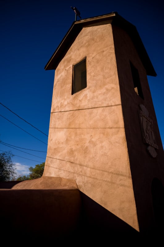 La torre de adobe color canela con dos ventanas y un escudo de armas proyecta fuertes sombras contra el cielo azul profundo. Las líneas eléctricas atraviesan la escena y los árboles son visibles al fondo.
