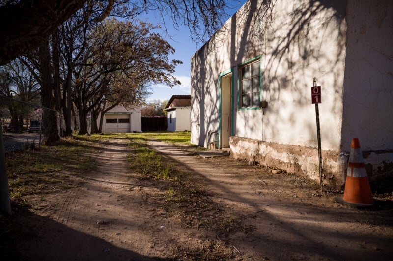 Un camino de tierra discurre a lo largo de un edificio blanco y desgastado con puertas y ventanas verdes. Las sombras de los árboles cercanos caen sobre la escena. Marcado con conos de tráfico y grifos. "01" Todo cerca del edificio.