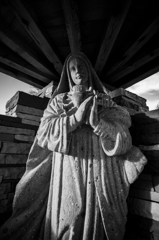 La fotografía en blanco y negro muestra una estatua de piedra de una mujer con una túnica con capucha, las manos entrelazadas sobre el pecho, de pie bajo un techo de madera con paredes de piedra apiladas al fondo.