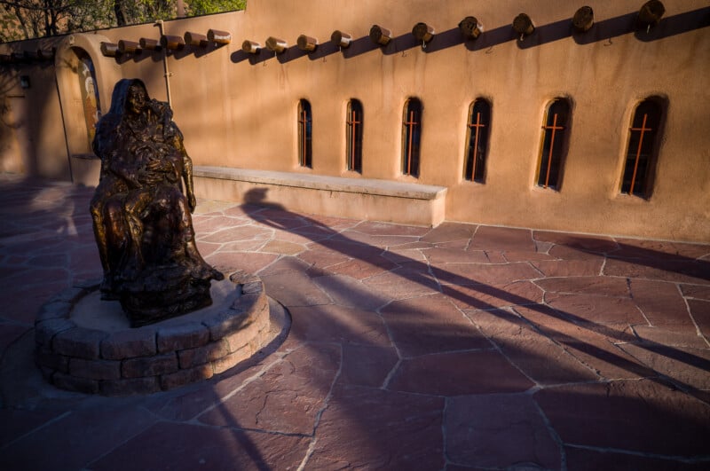 Una estatua de bronce se asienta sobre un pedestal de piedra en un patio soleado con azulejos de piedra roja. Largas sombras se extienden sobre el suelo, sobre un fondo de paredes de estuco con pequeñas ventanas y vigas de madera que sobresalen.