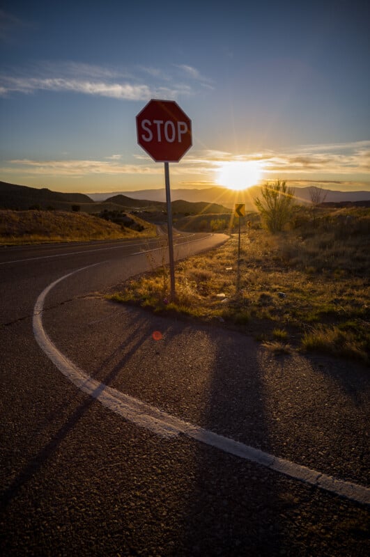 Al atardecer, una señal de alto se encuentra a lo largo de la carretera con curvas, proyectando largas sombras sobre la acera. Al fondo aparecen colinas y montañas bajo un cielo parcialmente nublado.