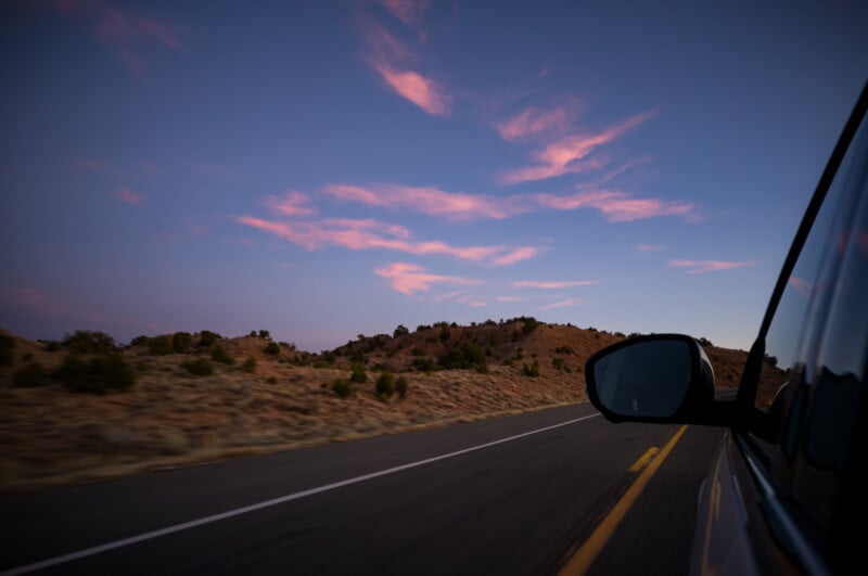 Un automóvil conduciendo por una carretera vacía al atardecer con nubes rosadas en el cielo azul con vistas al paisaje desértico y las colinas. En primer plano se pueden ver los espejos laterales y las ventanillas del vehículo.