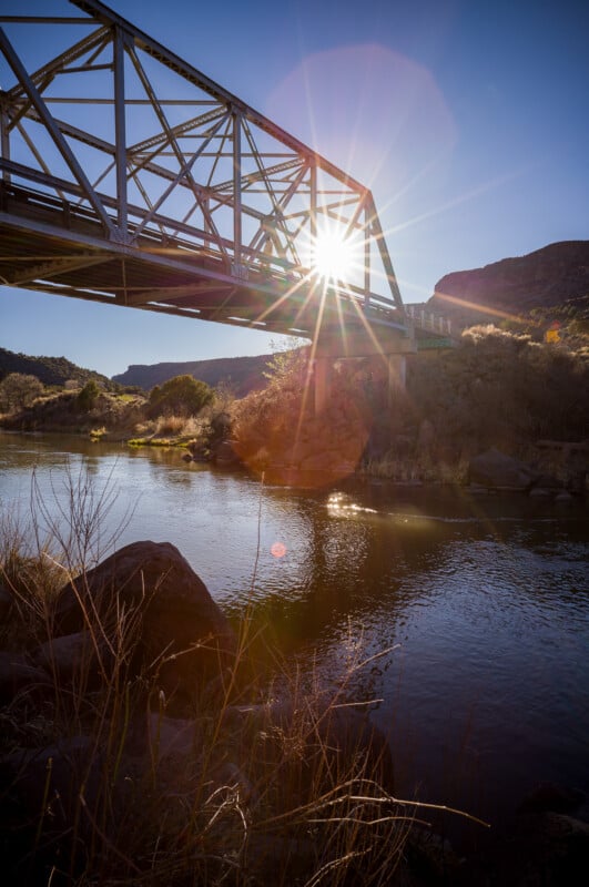 Un puente de metal cruza el tranquilo río y la luz del sol brilla a través del puente, creando un efecto similar al de un estallido de estrella. Bajo un cielo azul claro, colinas rocosas y escasa vegetación bordean las orillas del río.